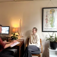A chiropractor at a desk talking with a woman seated opposite, a model spine on the desk and a nervous system chart on the wall