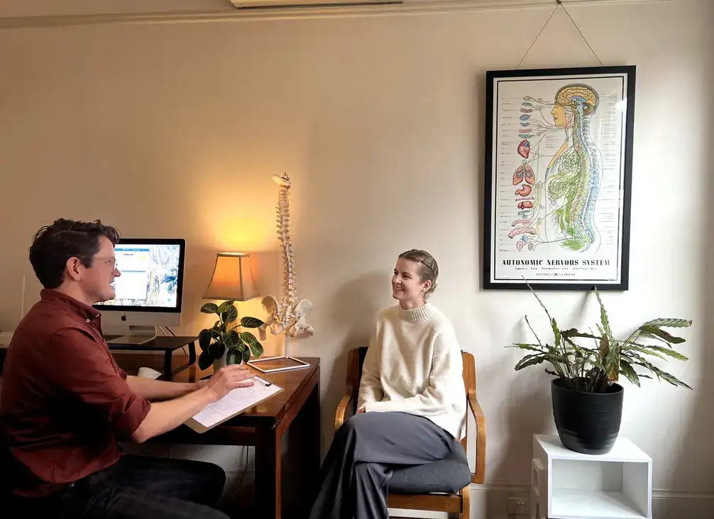 A chiropractor at a desk talking through a treatment plan with a patient, with a spine model beside them