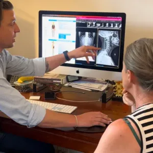 A chiropractor pointing to spinal X-rays on a computer screen while a woman seated beside him looks on
