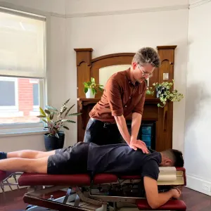 A chiropractor in a brown shirt with both hands on the mid back of a man lying face down on a treatment table